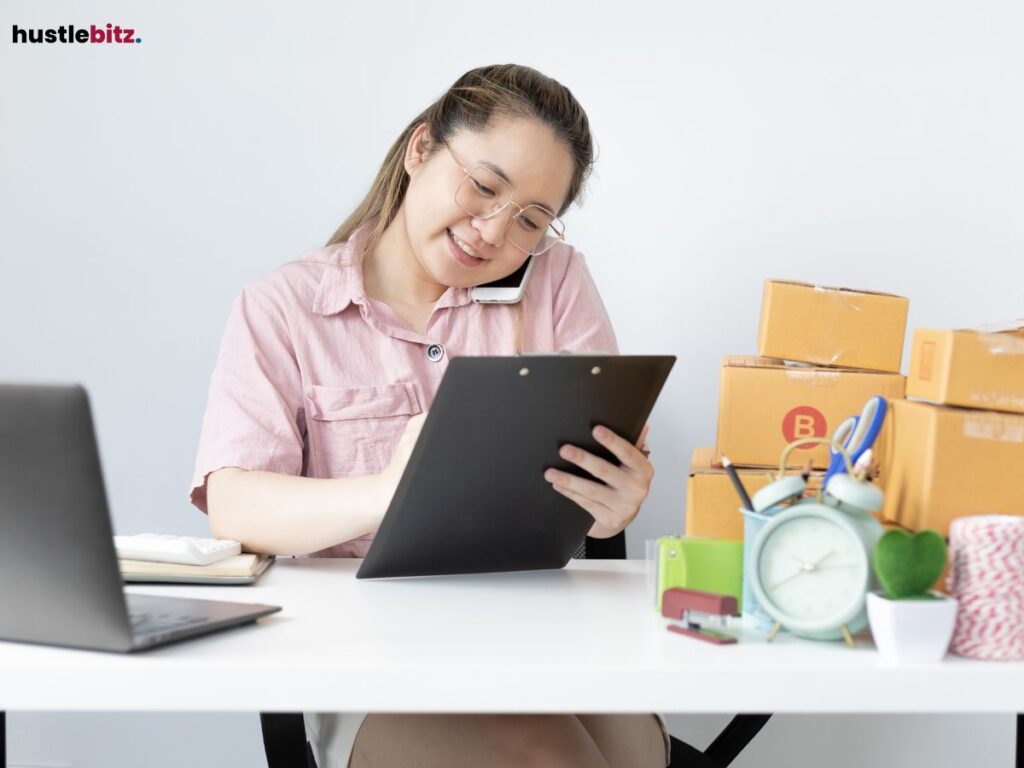 Woman talking on the phone while writing, surrounded by office supplies.