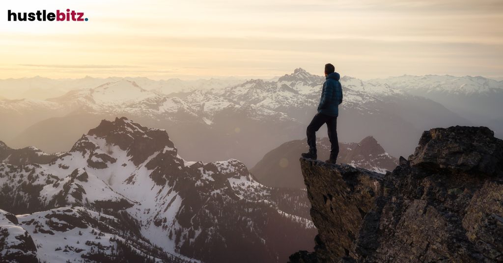 A person stands on a cliff, overlooking a vast mountain range at sunset.