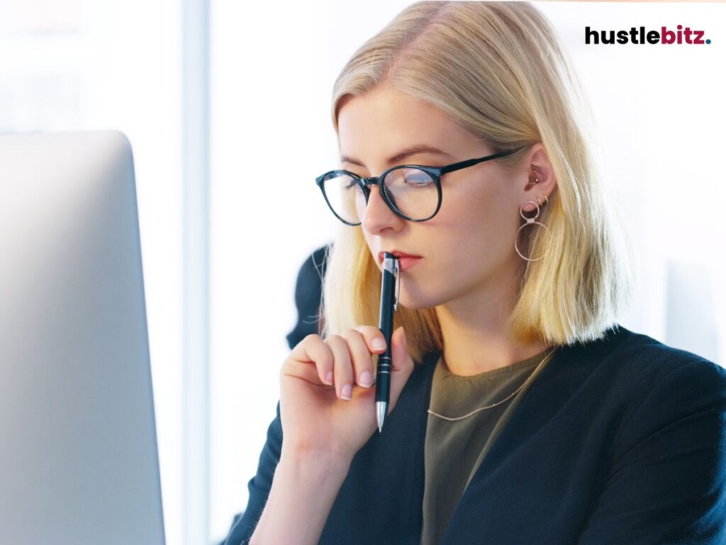 A woman in glasses, thoughtfully holding a pen while looking at a computer screen.