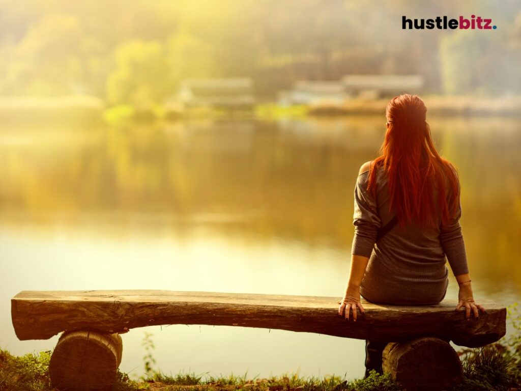A woman sits on a wooden bench, gazing peacefully at a calm lake in nature.