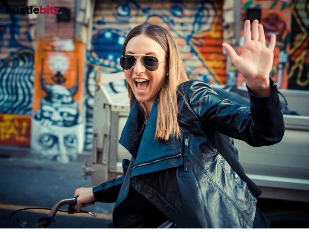A woman riding a bike wearing eyeglass smiles and raise her left hands