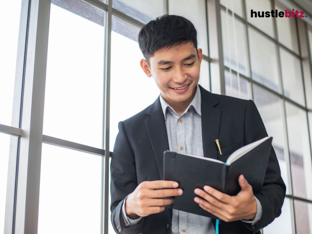 A man in a suit smiles while reading a notebook by the window.