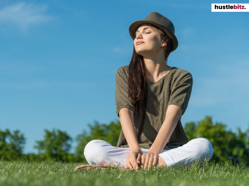 Young woman sitting cross-legged on grass, enjoying the sunlight with a hat.
