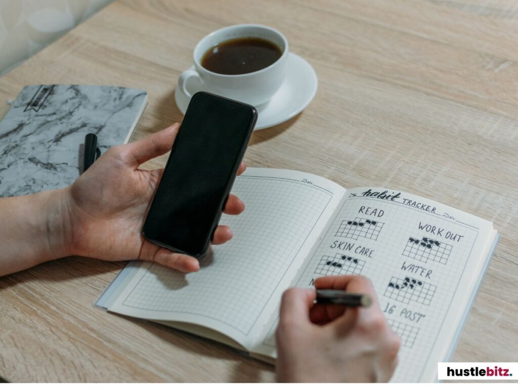 A person using a habit tracker in a notebook, with a phone in hand and a cup of coffee nearby.
