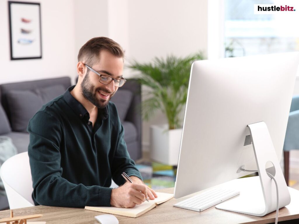Man smiling while writing notes at a desk, with a computer in front of him