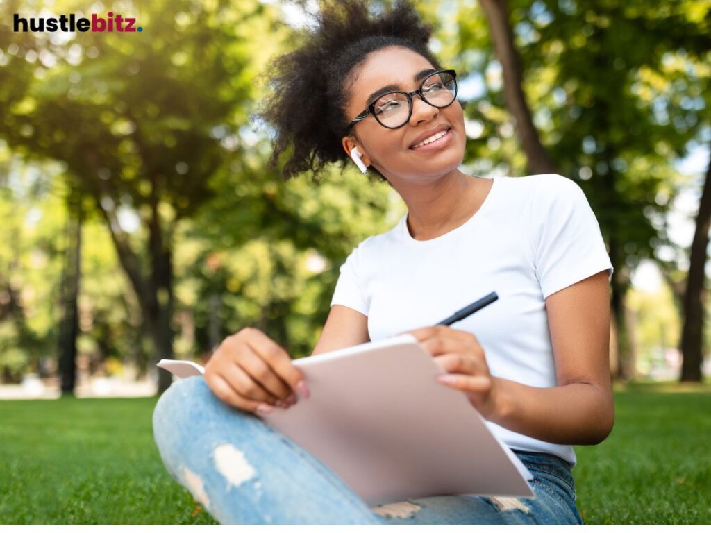 A young woman sits outdoors, writing in a notebook with a joyful expression.