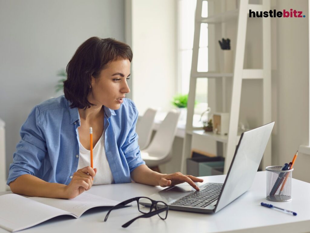 A focused woman works at a desk with a laptop, holding a pencil thoughtfully.