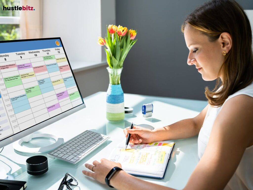 A woman writing in a planner beside a monitor displaying a colorful calendar.