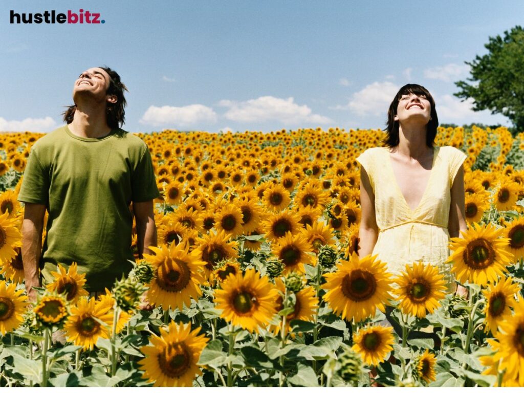 Two people stand smiling in a bright field of sunflowers, facing the sky.