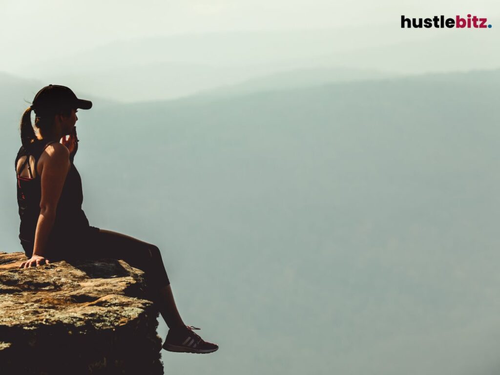 A woman sits on a cliff, gazing at misty mountains, reflecting on the view.