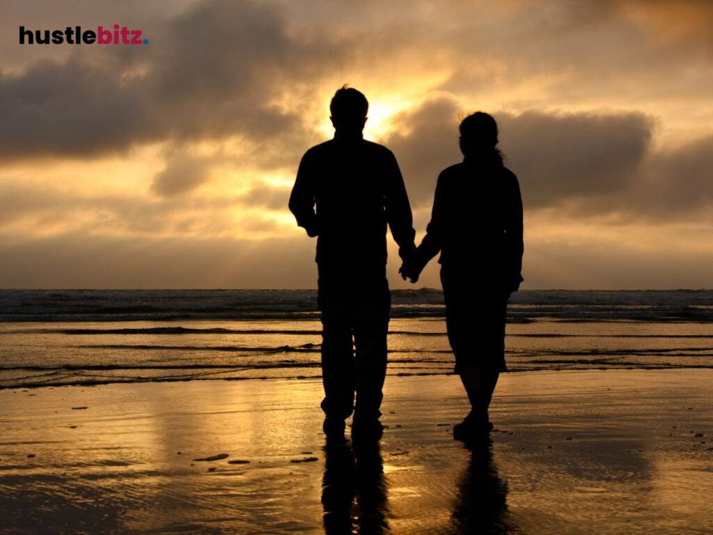 A couple walks hand in hand on the beach at sunset, their silhouettes glowing.
