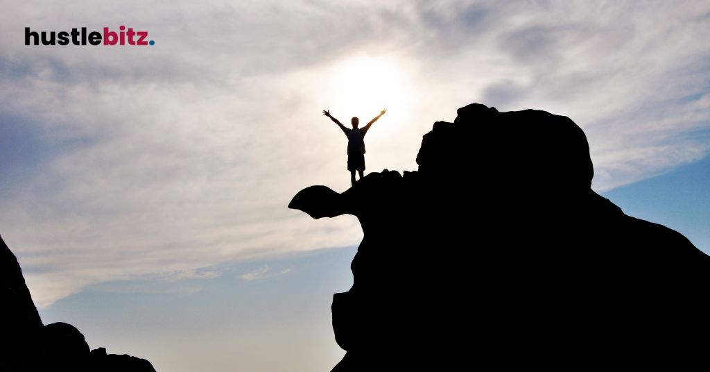 A person stands on a rocky peak, arms raised in triumph against a bright sky.