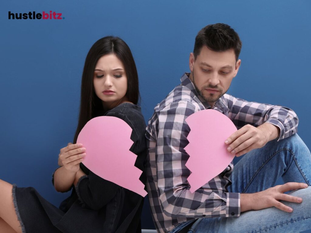 A couple sits back-to-back holding two halves of a broken pink heart.