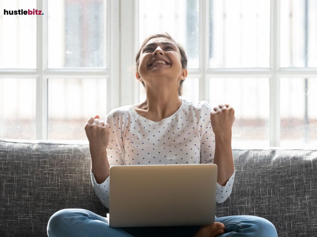 Woman celebrating with fists raised, sitting on a sofa with a laptop.
