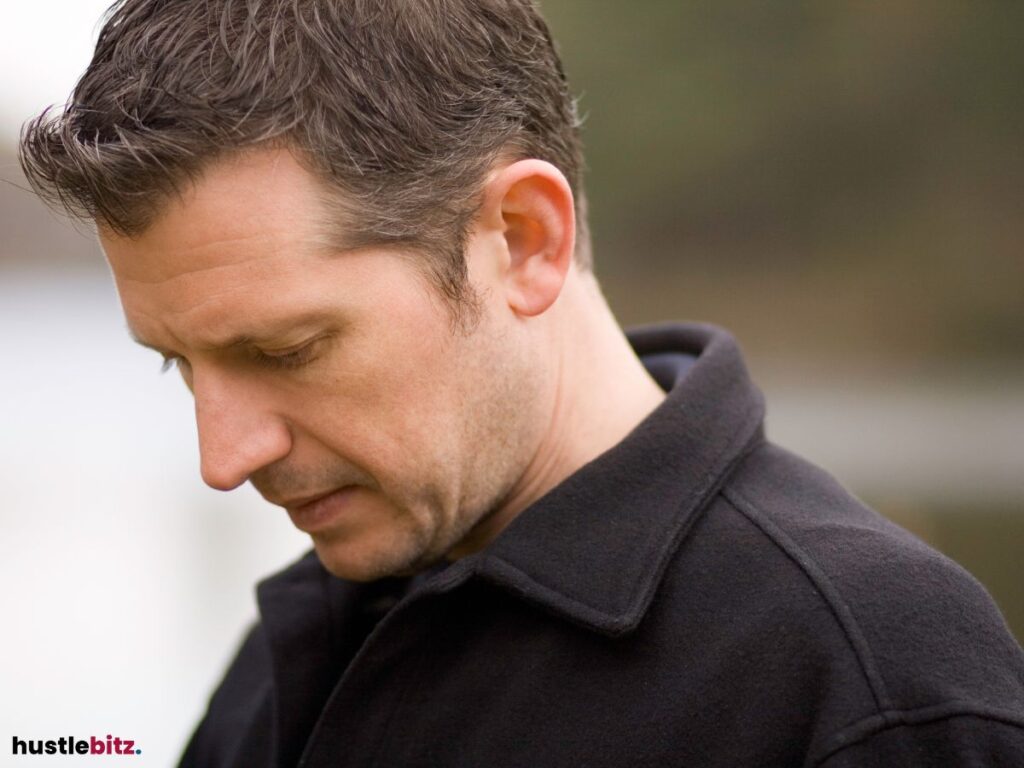 Man looking down thoughtfully, dressed in a dark coat.