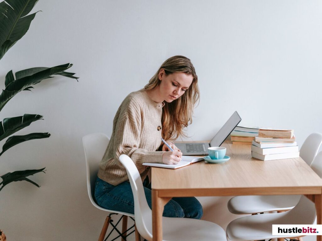 A woman writing in a notebook while working on her laptop at a desk.