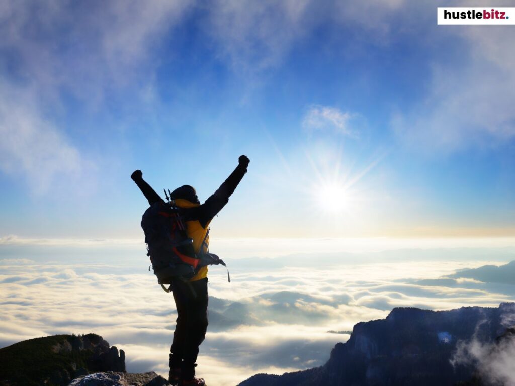 A hiker on top of a mountain, raising arms in triumph under a clear sky.