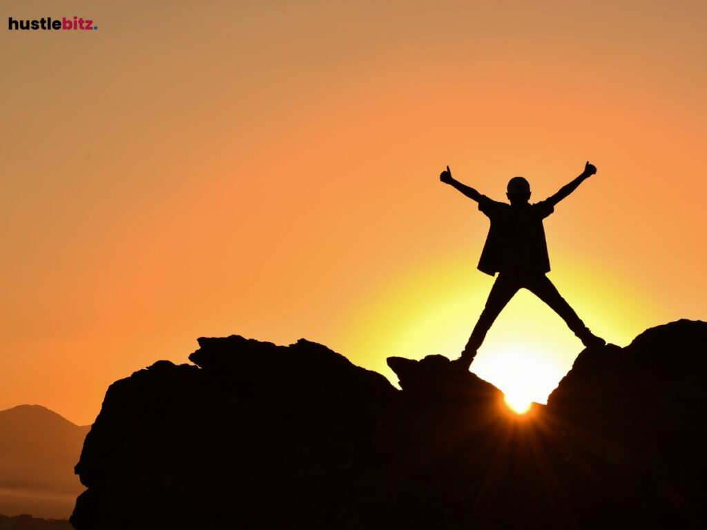 A silhouette of a person standing triumphantly on rocks against a sunset background.