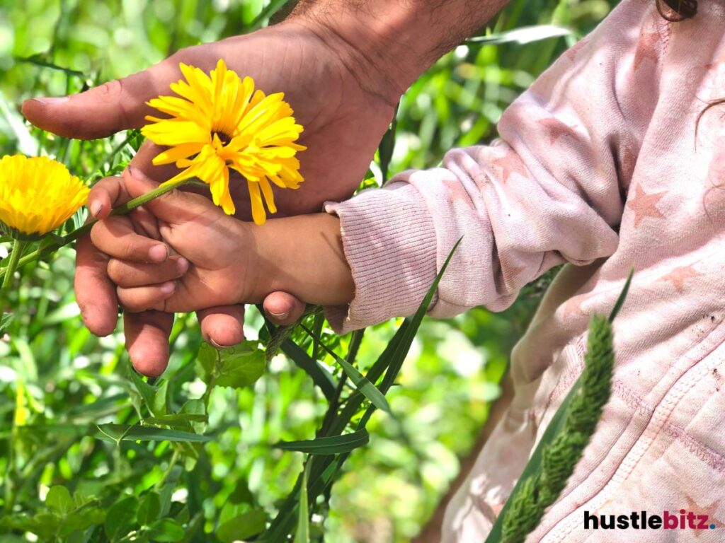 Adult and child holding hands while picking yellow flowers in a garden.