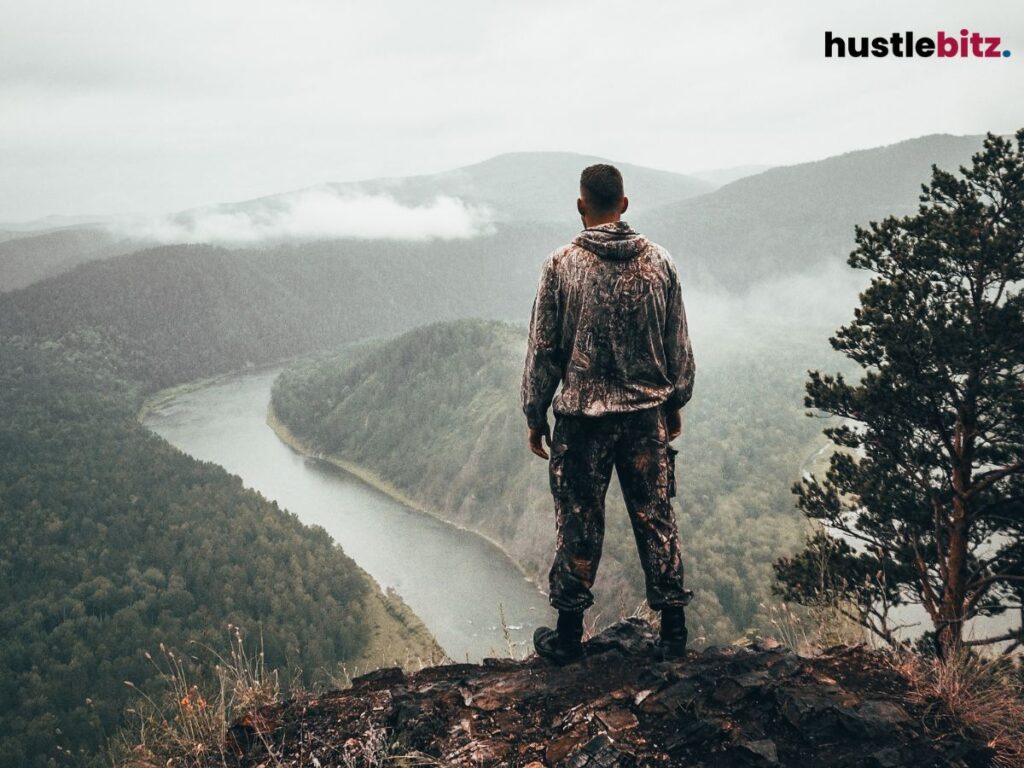 Man standing on a cliff, overlooking a winding river and a forested valley.